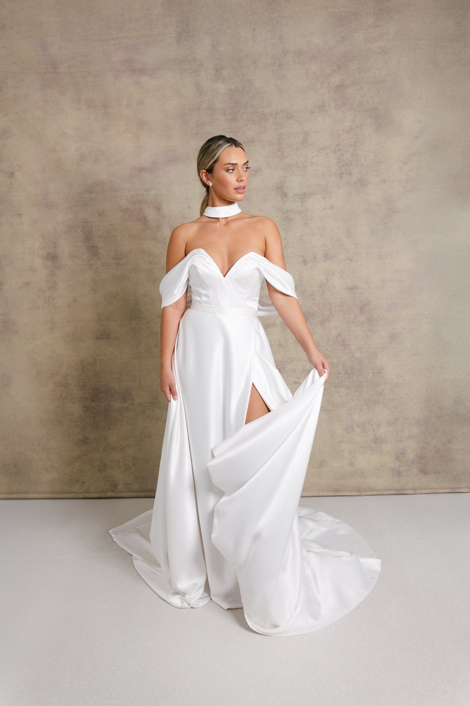 A woman in a luxurious slipper satin two piece wedding dress, featuring a sweetheart corset top with off the shoulder bardot sleeves and slipper satin wrap over leg split skirt.