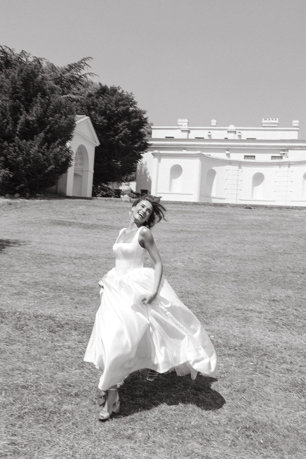 Woman in a ivory square neckline wedding dress standing on grass with a building and trees in the background