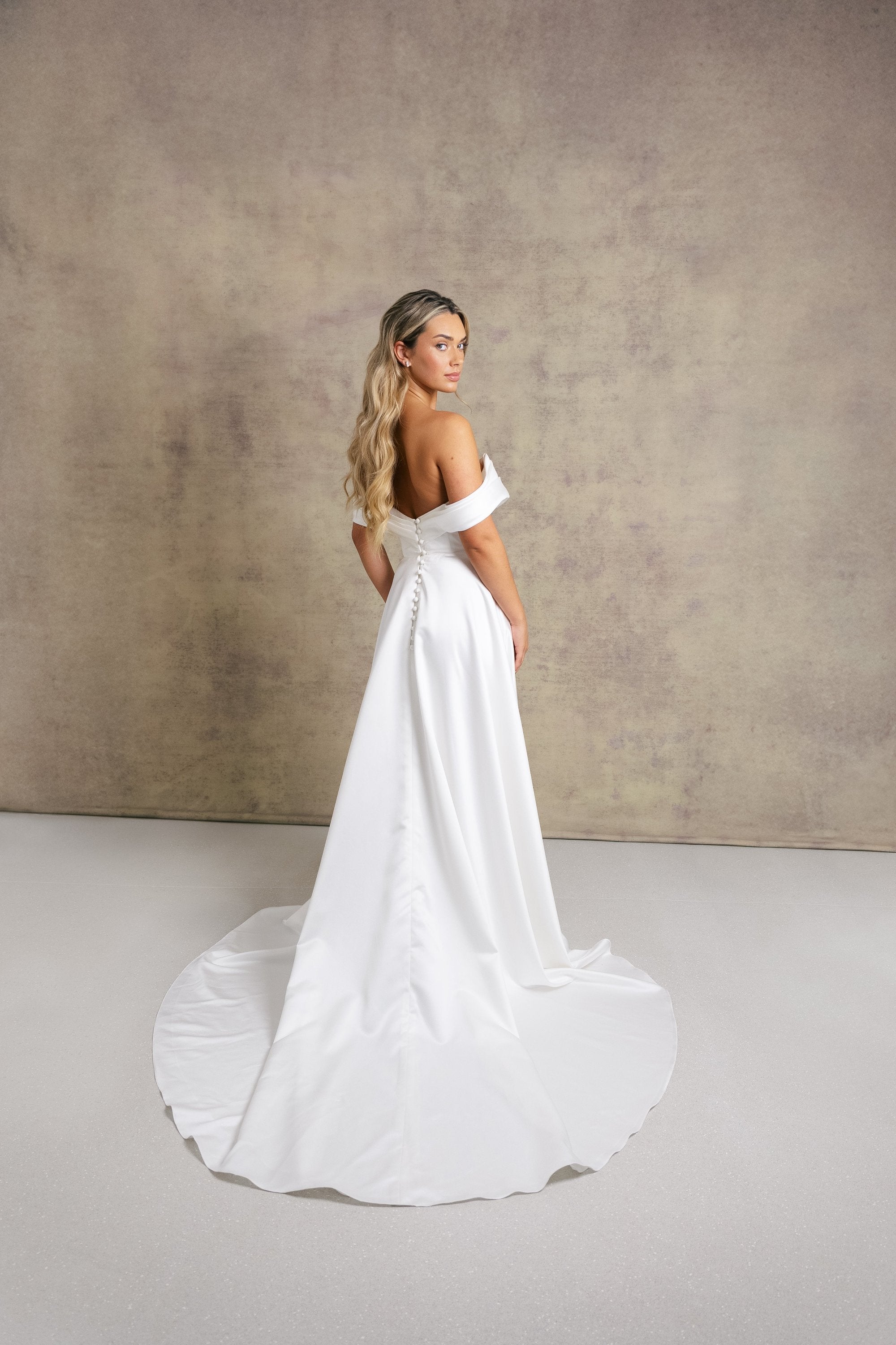 A woman in a subtle dropped waist wedding dress, off the shoulder boned bodice with a soft draped neckline and a wrap over leg split skirt in our gorgeous satin fabric.