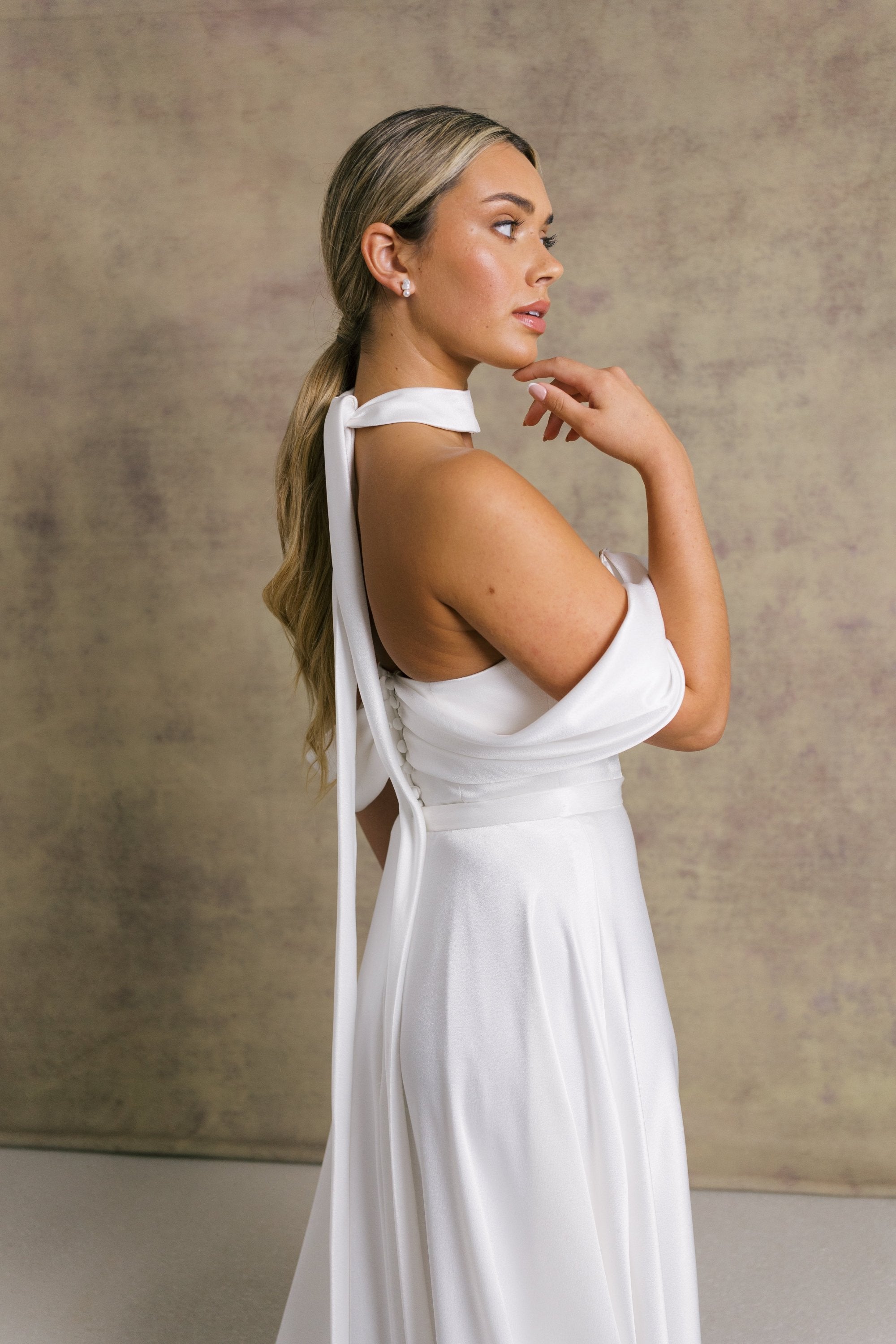 A woman in a luxurious slipper satin two piece wedding dress, featuring a sweetheart corset top with off the shoulder bardot sleeves and slipper satin wrap over leg split skirt.