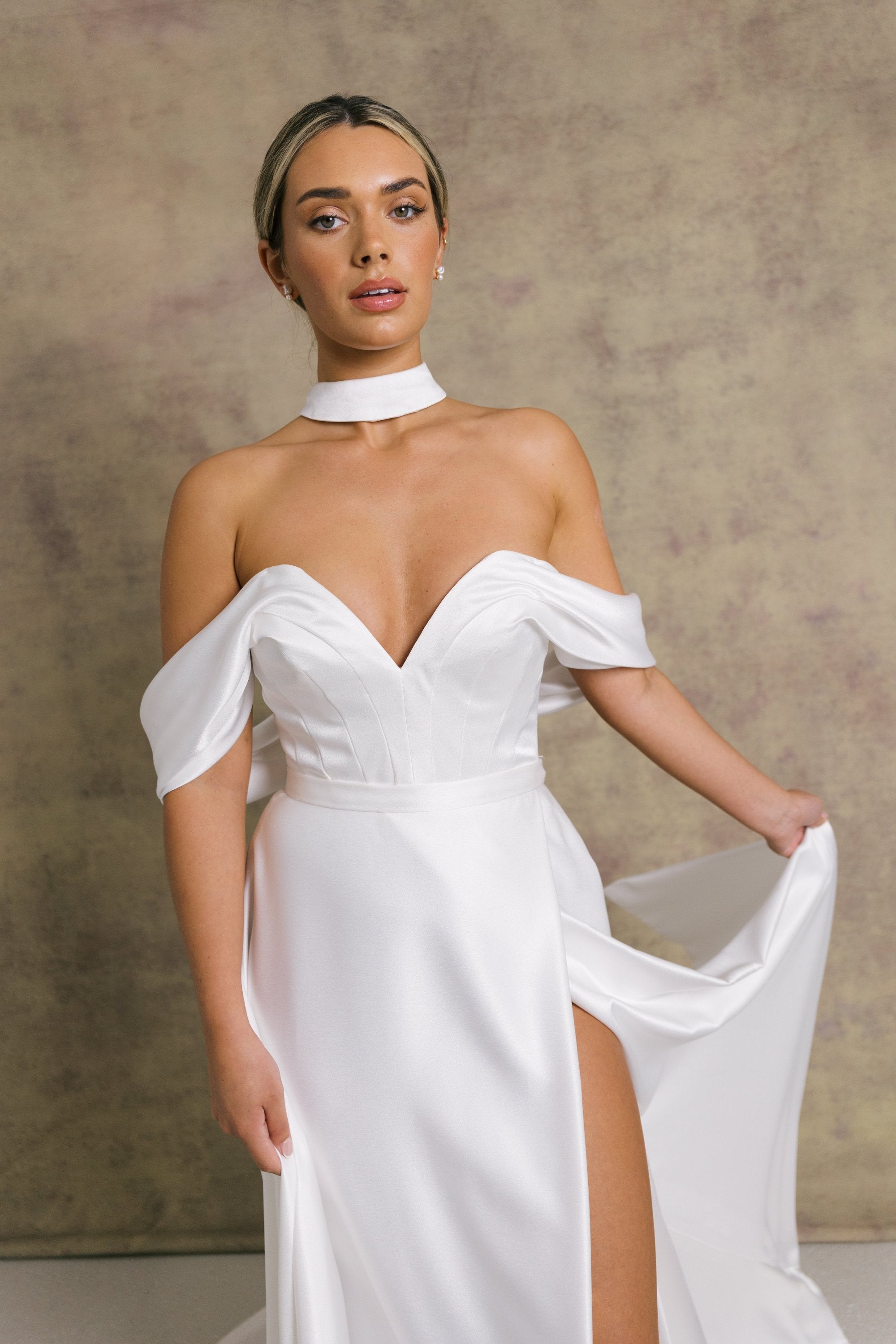 A woman in a luxurious slipper satin two piece wedding dress, featuring a sweetheart corset top with off the shoulder bardot sleeves and slipper satin wrap over leg split skirt.