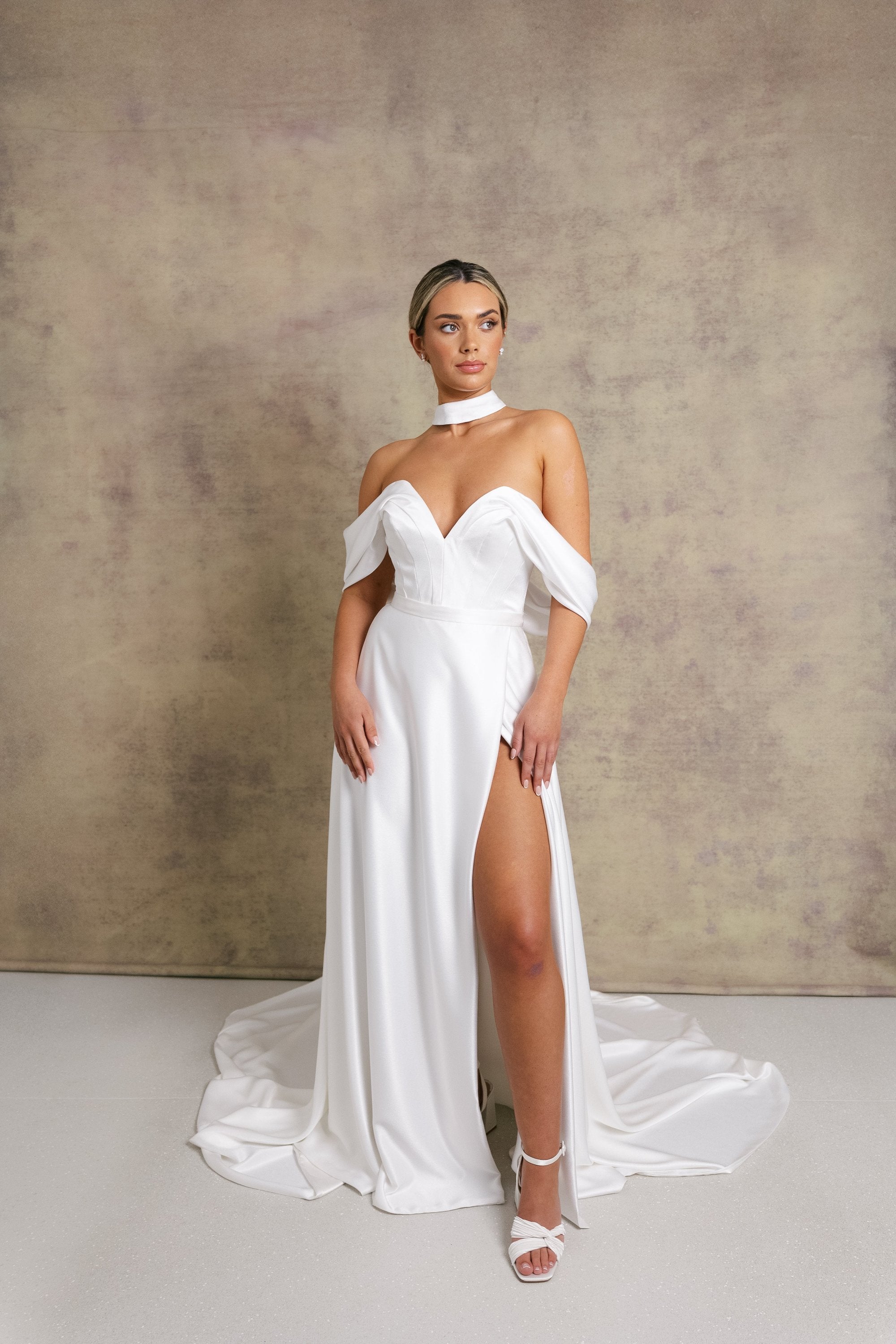 A woman in a luxurious slipper satin two piece wedding dress, featuring a sweetheart corset top with off the shoulder bardot sleeves and slipper satin wrap over leg split skirt.