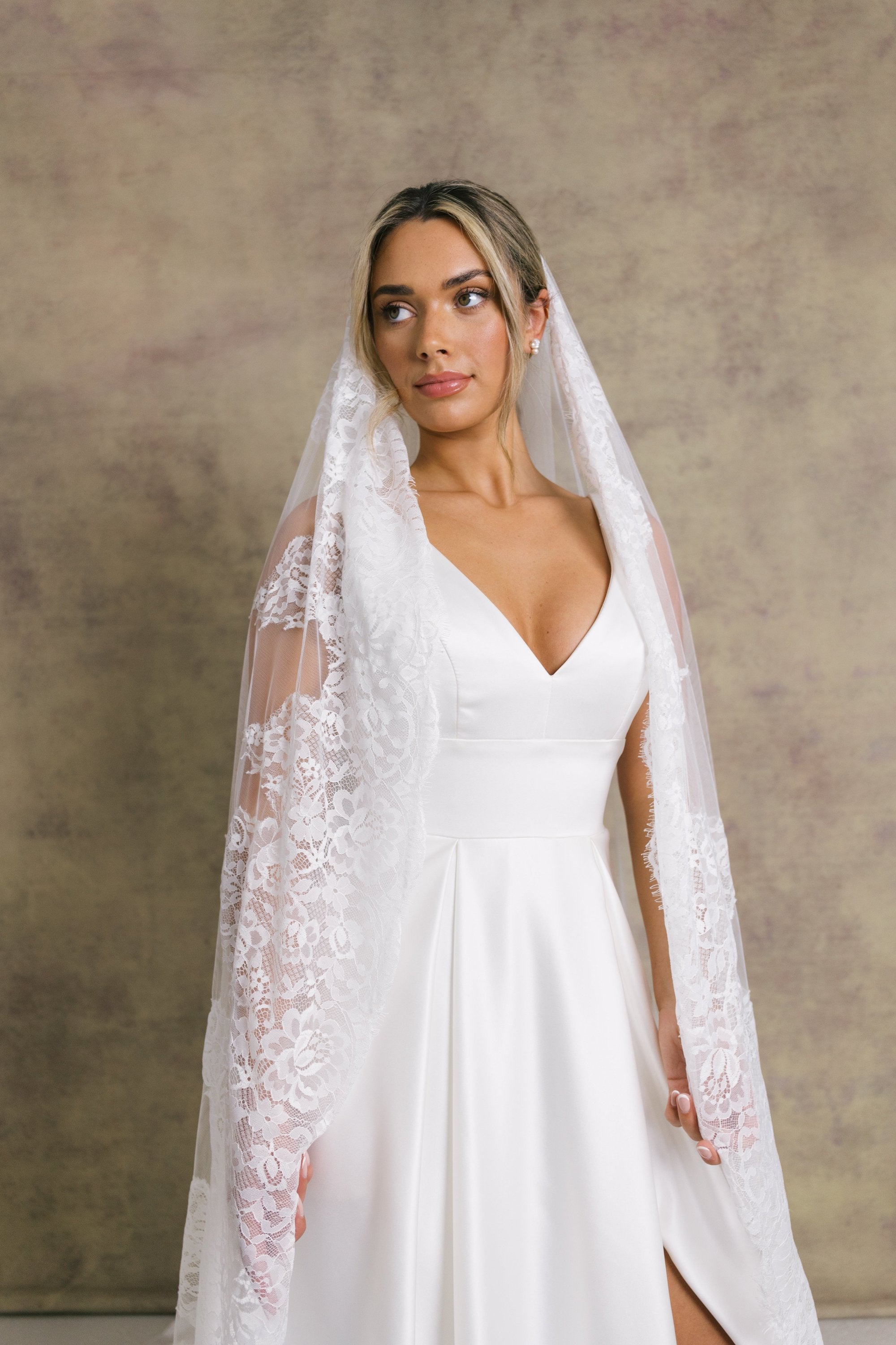 A woman in a striking v neckline wedding dress with waistband detail on a box pleated satin skirt, complemented with a Chantilly lace border veil.