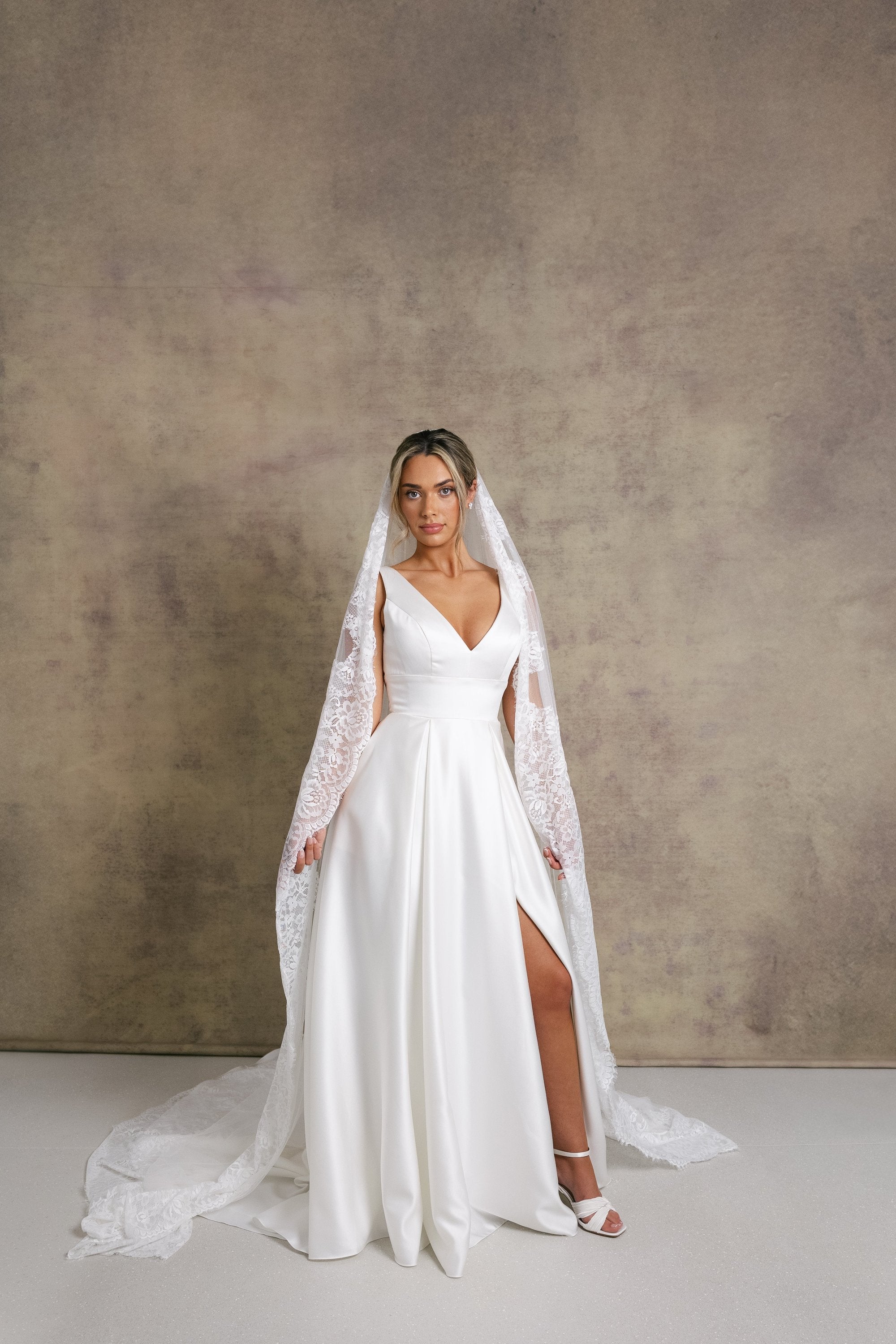 A woman in a striking v neckline wedding dress with waistband detail on a box pleated satin skirt, complemented with a Chantilly lace border veil.