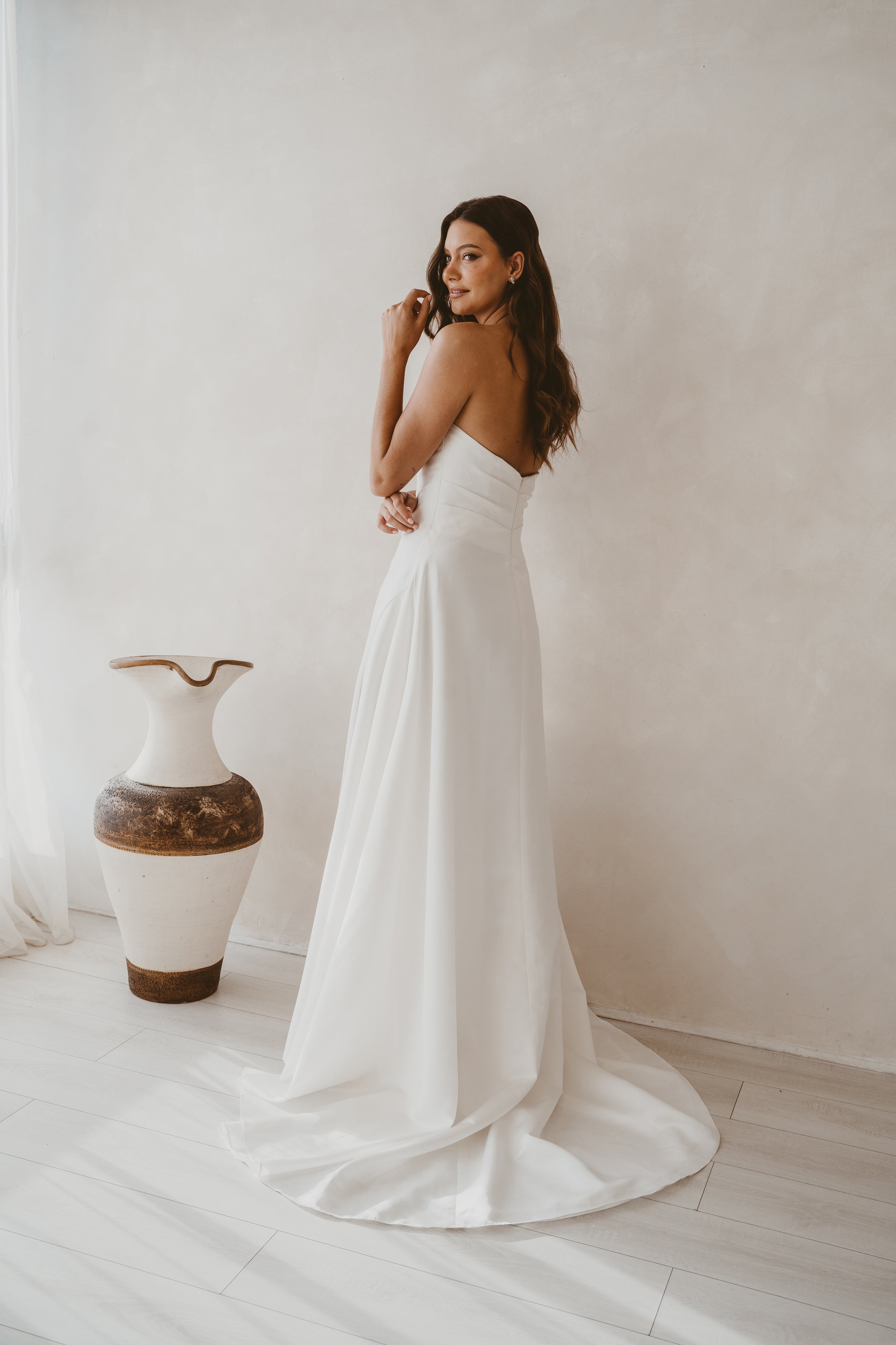 Woman in a white dress standing against a plain wall with a decorative vase in the background.